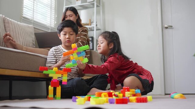Asian children playing with colorful building blocks while their mother works from home on a laptop in the living room