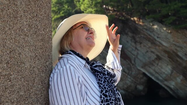 Mature Woman Enjoying Seaside Vacation. Mature woman in sun hat and sunglasses smiling by the sea with rocky coast in the background during summer vacation.