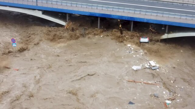 Aerial view of a car being swept away and violently thrown against a bridge by a river overflowing due to flooding.