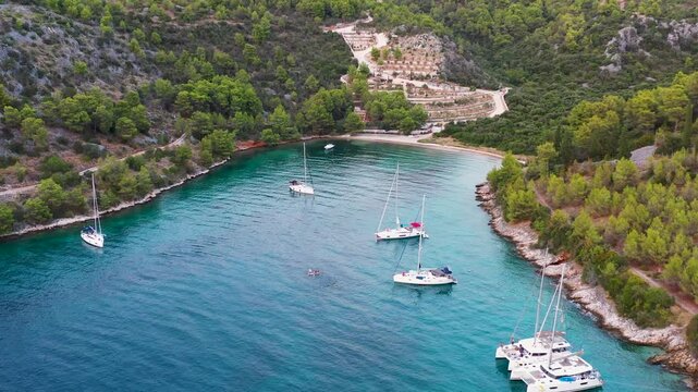 A pebble beach closes the head of Uvala Stiniva near Sutivan on the island of Brac, Croatia, charter yachts at anchor below dry stone terraces and pine forest. Aerial rising shot