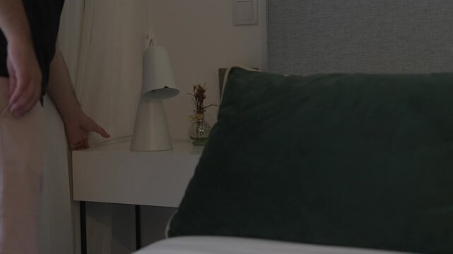 Person's hand reaching to turn on or off a modern lamp on a bedside table in a cozy bedroom setting, showing home decor