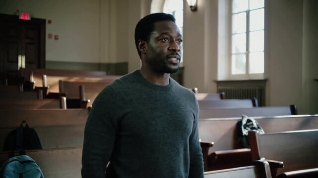 Making gesture, man in dark sweater standing on classroom bench with backpacks, speaking to address