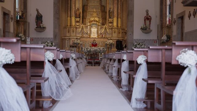 Church aisle and pews adorned with white flowers and tulle, set for an upcoming wedding ceremony. Golden altar in background