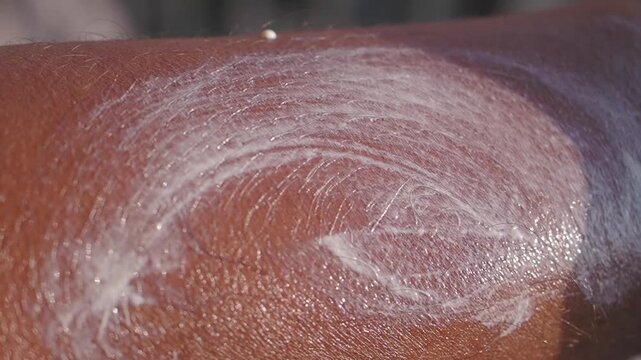 Close up of a person applying white lotion or cream onto dark skin, creating circular patterns that slowly blend as it is rubbed in