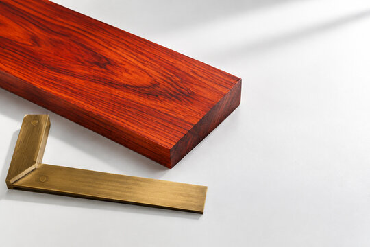 Polished padauk wood slab with brass carpenter's square, bright studio surface, wide crop