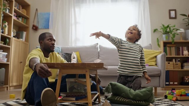 African American father and young son throwing paper airplane while sitting on floor at home. Perfect for family lifestyle commercials, parenting campaigns, and fatherhood storytelling