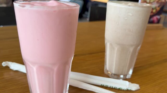 Overhead top-down view looking directly into the two milkshake glasses, capturing the swirled pink surface and frothy cream top alongside crossed straws on the wooden table below