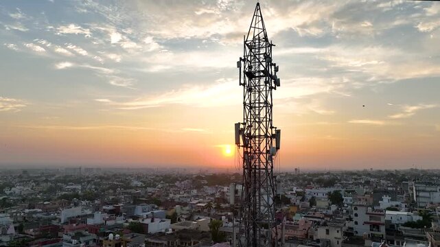 Cinematic circling drone view of high-voltage telecommunications mast and microwave dishes above urban sprawl.