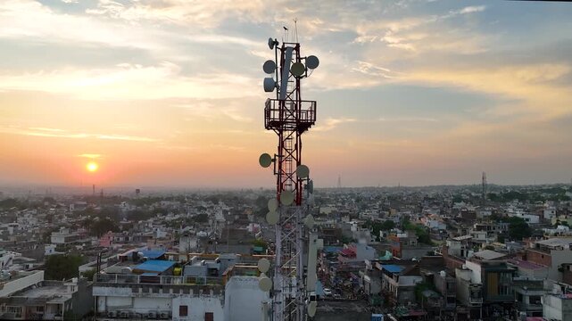 Aerial circling motion around high-voltage telecom tower antennas overlooking a dense residential Indian settlement.
