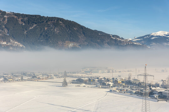 Snowy Kaprun valley lies beneath forested Austrian Alps, wrapped in low winter fog and bright sunlight. Power pylons cross frozen fields near village, roads, farms, and industrial buildings