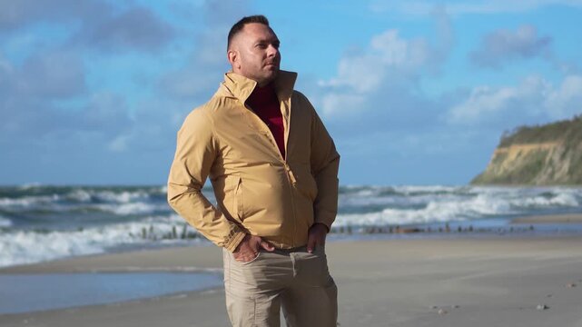Smiling handsome man in a beige jacket posing on a sandy beach of the Baltic Sea with a static camera shot.