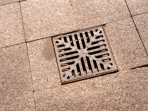 Close-up of a square metal storm drain grate installed in a paved pedestrian walkway. Urban infrastructure detail representing drainage, rainwater management, sewer systems, street engineering and pub