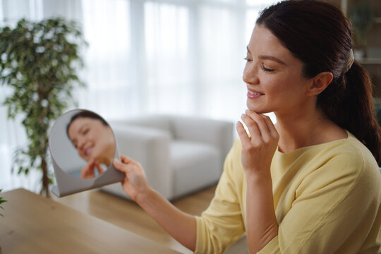 Woman holding a small mirror checking her smooth facial skin, enjoying a morning self-care beauty routine at home, promoting health and wellness