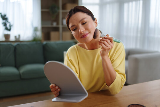 Woman applying face powder with a makeup brush, looking into a small mirror, performing her daily beauty routine and self-care at home