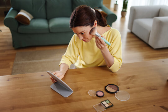 Woman applying face makeup using a brush and mirror, creating her beauty routine at home, enhancing appearance and practicing self-care for skin grooming
