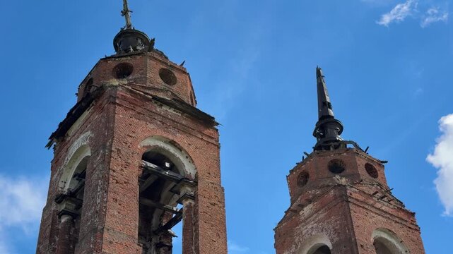 Two ancient brick bell towers with arched openings, dilapidated masonry, and striking spires stand against the backdrop of a bright blue sky with light clouds.