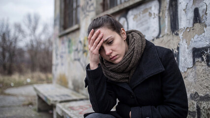 Fototapeta premium Depressed woman sitting outdoors near a dilapidated building touching her forehead in deep sorrow.