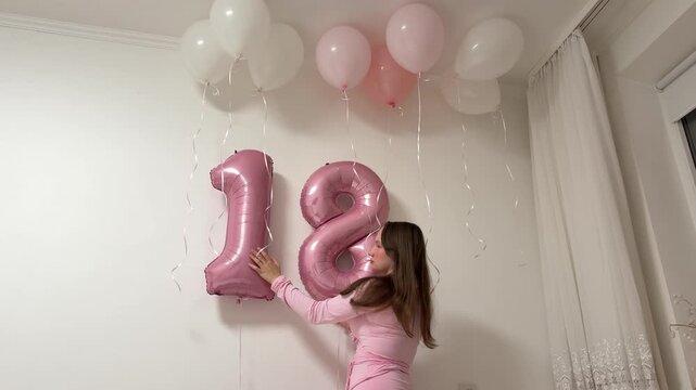 Creating a festive birthday backdrop, a young woman arranges balloons and carefully adjusts the number 18 decoration on the wall for her coming-of-age celebration.