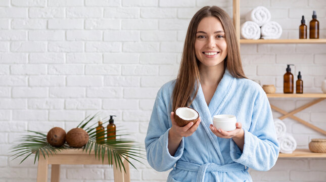 Woman in blue bathrobe holds coconut and cream jar in spa setting with shelves of skincare products and towels in the background