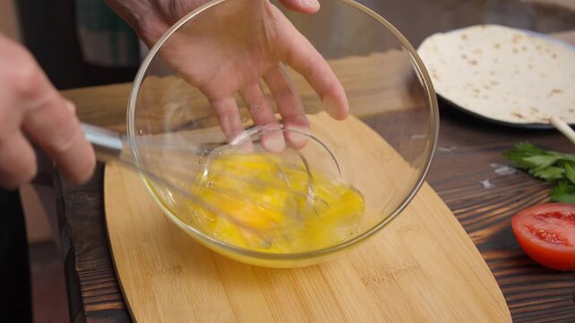 Close-up of a chef whisking raw eggs in a glass bowl with a metal wire whisk in 4K. Preparation of ingredients on a wooden board for cooking breakfast, culinary and food concept.