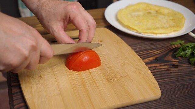 Close-up of a chef slicing a fresh red tomato with a kitchen knife on a wooden cutting board in 4K. Preparing ingredients for breakfast or wrap, culinary and healthy food concept.