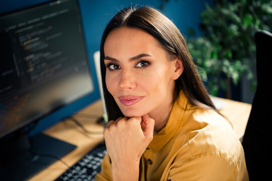 Confident female software developer looking at camera in a modern office workspace with monitors and keyboard, portrait of a professional coder at her desk