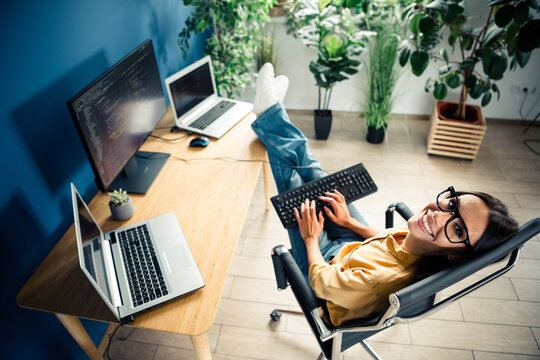 Smiling female developer relaxing in a modern home office with laptop, monitor and keyboard while coding and working remotely in a bright, plant-filled workspace