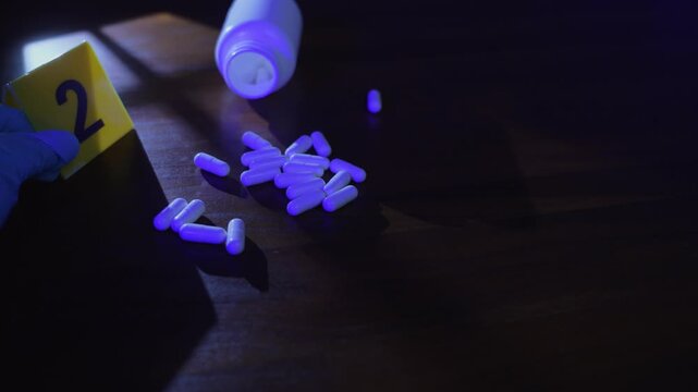 Crime scene investigation of a gloved hand placing a yellow evidence marker next to scattered white pills and a bottle on a wooden table under window frame shadows and police lights.