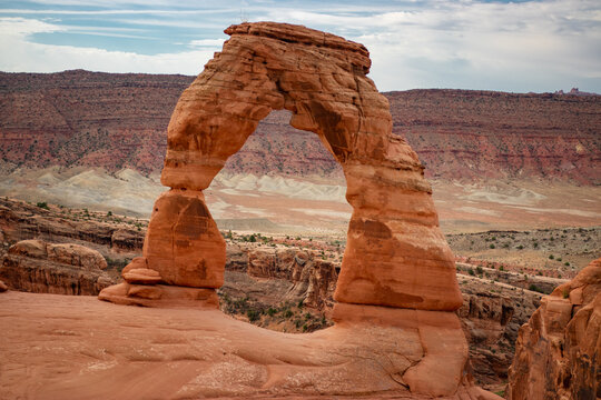 View of Delicate Arch in Arches National Park, featuring iconic red sandstone rock formation against a desert landscape with distant mesas in Grand County, Utah, United States.