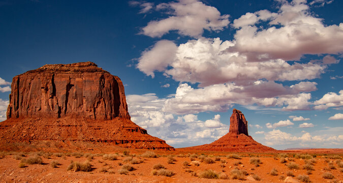 View of the iconic red sandstone buttes rising from the desert floor under a bright blue sky with scattered white clouds in Oljato-Monument Valley, Arizona, United States.