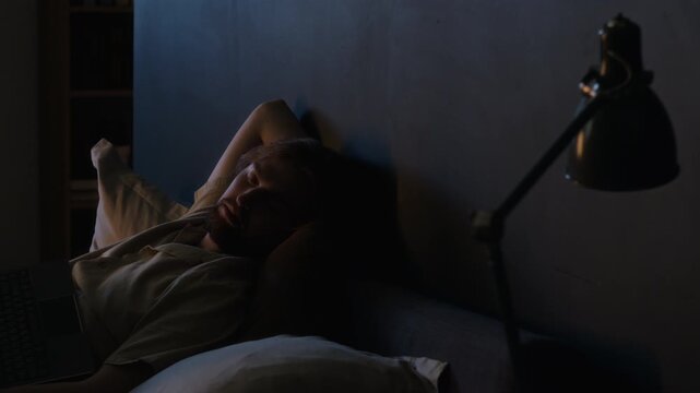 Young adult Caucasian man sleeping in bed beside laptop after working late at night. Footage portrays digital fatigue, insomnia, and remote work burnout for healthcare or lifestyle campaigns