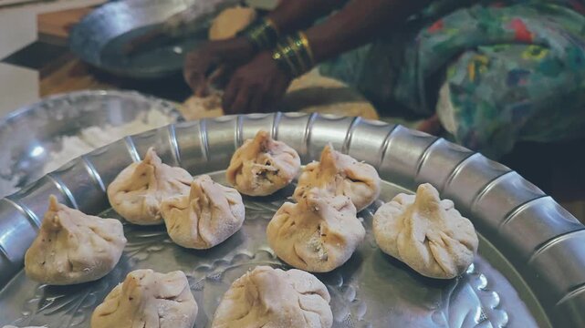 Intricately pleated modak dumplings sit ready to steam on a silver tray while a woman continues the sacred, ancestral craft of handshaping sweet offerings on the kitchen floor for the Ganesha festival