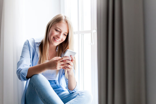 Happy woman using smartphone at home, sitting by window. Concept of shopping online, distance learning, banking, finance. Freelancer working, relaxing. People connection, communication. Copy space
