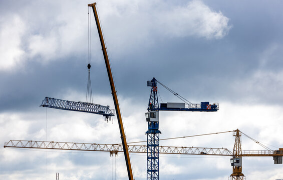View of construction cranes working on a building site with a metal truss suspended in the air against a cloudy sky in Vilnius, Vilnius County, Lithuania.