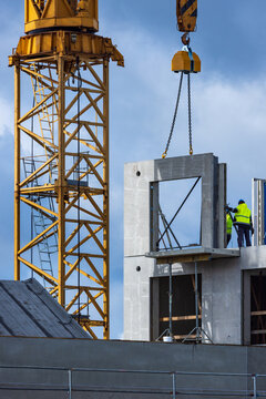 View of a yellow tower crane lifting a concrete wall panel at a construction site with workers in high-visibility vests in Vilnius, Vilnius County, Lithuania.