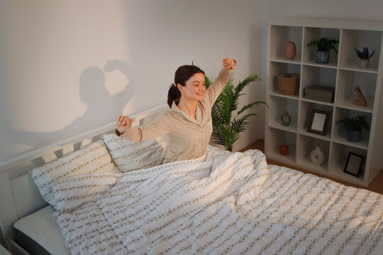 Young woman waking up in bed, happily stretching arms after good night's sleep, embracing fresh morning light and feeling refreshed in a cozy bedroom setting