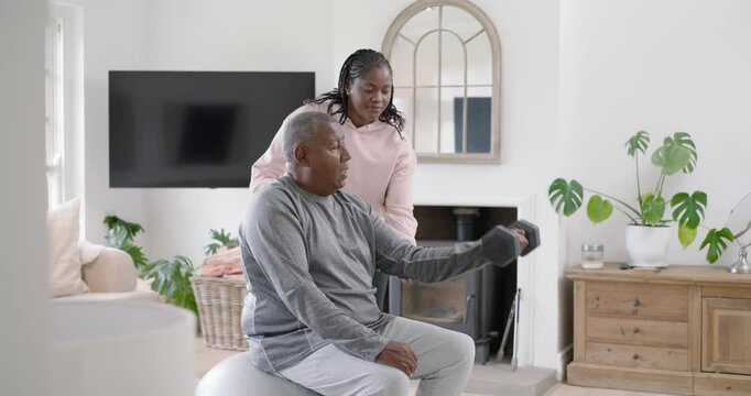 Caregiver in hoodie offering hex dumbbell, senior man on exercise ball taking curls for home rehab