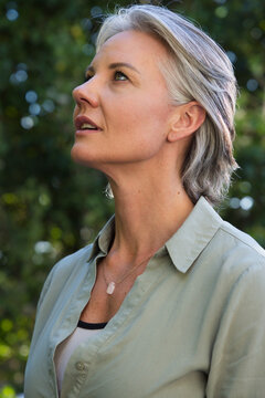 Senior woman looking upward in garden, wearing green button shirt, ribbed tank and pale pendant