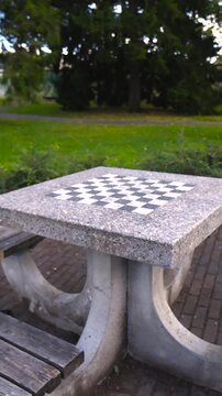 Granite chessboard table with attached wooden benches sitting empty in a lush green public park, offering a quiet spot for intellectual games, strategic thinking, and outdoor leisure