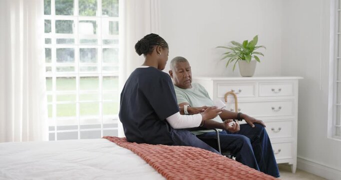 African American aide in scrubs placing cuff showing meter in bedroom for home visit, man watching