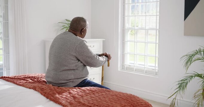 Senior African man sitting on bed in gray knit holding cane, sun lighting window, gazing outside
