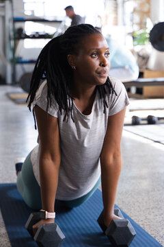 African woman kneeling on blue mat in gym gripping black hex dumbbells, wearing teal leggings