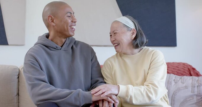 Senior Asian mother and man touching hands with his resting on knee, laughing together on couch