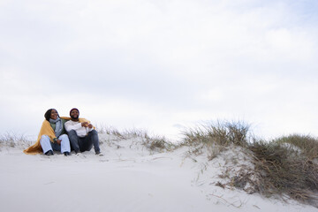 Obraz premium Couple sitting on sand dune at beach wrapped in yellow blanket wearing burgundy beanie, copy space