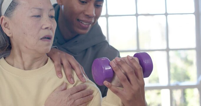 Senior Chinese woman holding purple weight African man touching shoulder guiding wrist PT by window