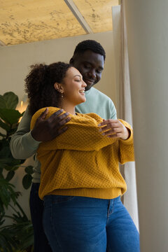African American couple embracing near tall window at home with potted plant, wearing knit sweater