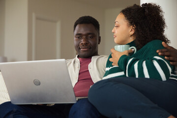 African American couple sitting on light sofa at home, sharing silver laptop and light blue mug © WavebreakMediaMicro