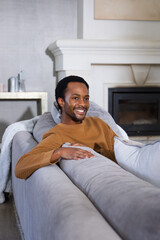 Obraz premium African American man reclining on gray sectional sofa in cozy living room, smiling in brown sweater