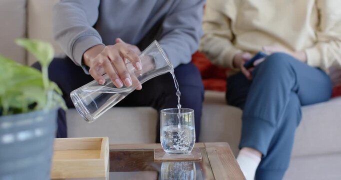 Pouring carafe, diverse duo on couch, man offering drink to Senior Asian woman holding phone caring
