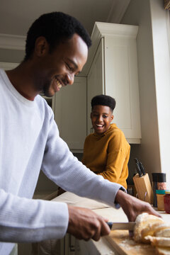 African father wearing grey top and teen son laughing at kitchen counter while father slicing bread